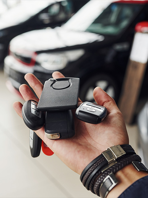 a person's hand holding multiple car keys and a key fob with a blurred background of cars in a dealership showcasing five unique key designs